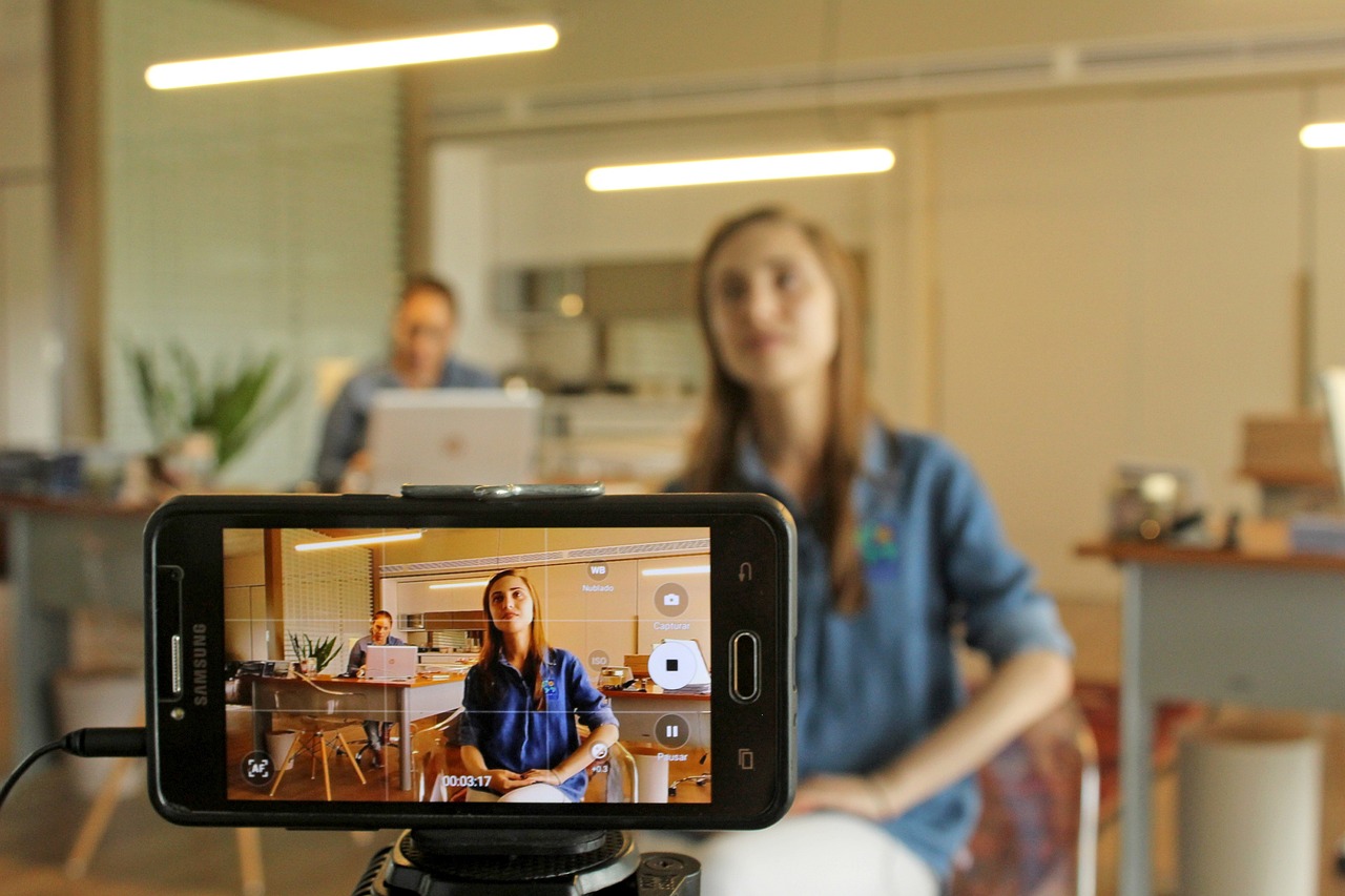 A yougn woman in front of a mobile phone camera on a tripod, recording herself.