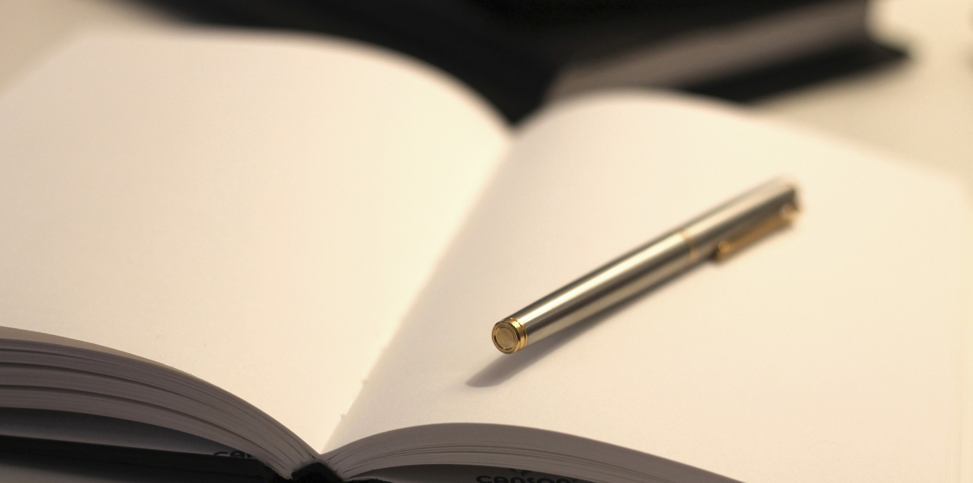 A blank notebook on a table with a silver coloured pen resting on the open page.