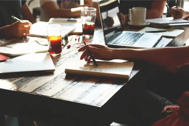 Several people sitting at at table with notebooks, laptops and drinks